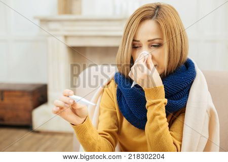 Sickness. Weak young blond woman feeling bad and blowing her nose and holding medication while having a blanket on her shoulders and sitting on the couch