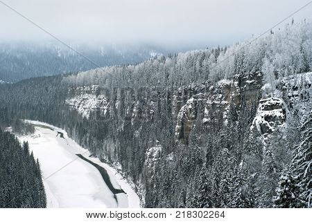 The view from the top of the rock to the frozen river flowing among the cliffs in the winter snow-covered gloomy landscape