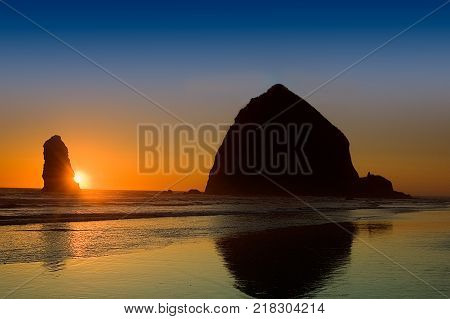 Haystack Rock Oregon reflected in surf with sun setting behind