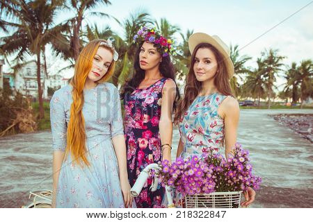 Beautiful women in a dresses stand near a bicycle on palm background.