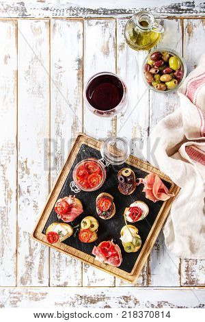 Tapas or bruschetta variety. Bread with ham prosciutto, sun dried tomatoes, olive oil, olives, pepper on slate wood serving board with glass of red wine over white wooden background. Top view, space