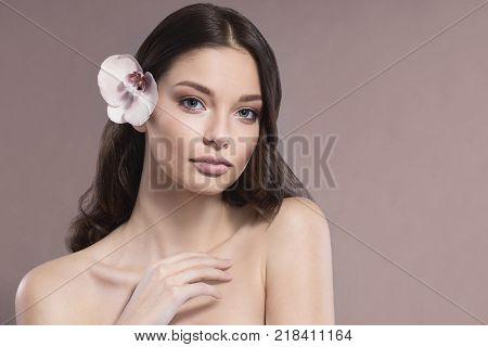 Young and healthy woman with light make-up on beige background. The hair is decorated with beautiful fresh Orchid flower. SPA and skin care concept.