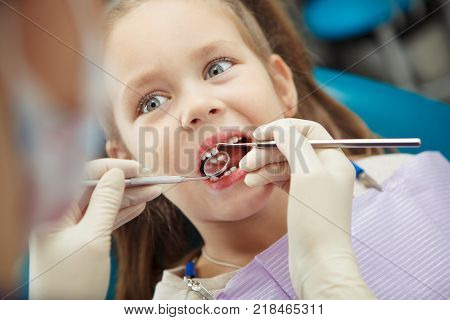 Child lies in comfortable dentis chair and goes through procedure without fear while female doctor in rubber gloves fix girls teeth in modern medical office.