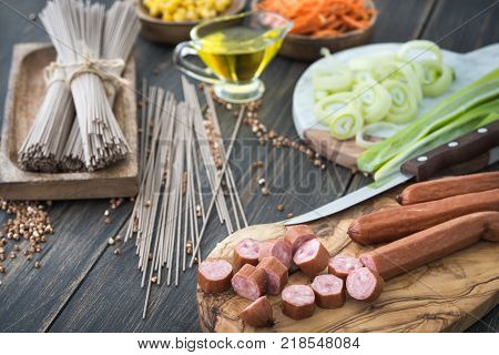 Slicing sousages for soba noodles cooking with leek, corn and korean carrot on a dark wooden rustic background