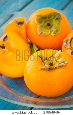Wet ripe persimmons or date-plum fruits in glass plate on blue rustic wooden background closeup