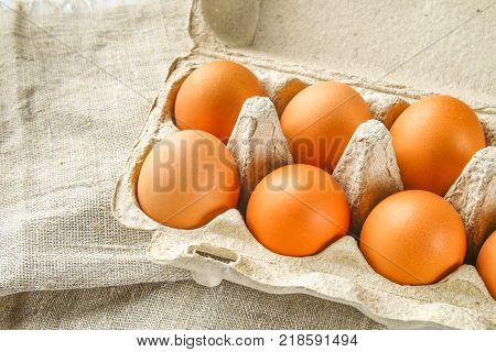 Raw brown chicken eggs in a cardboard tray with cells on sacking on a white wooden table. Ingredients for cooking