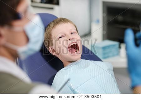 Male dentist examines the teeth of the patient cheerful child with blond hair. Boy smiling in dentist's chair with mouth wide open.