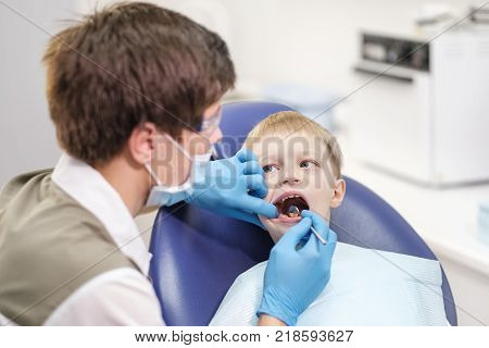 Male dentist examines the teeth of the patient cheerful child with blond hair. Boy smiling in dentist's chair with mouth wide open.