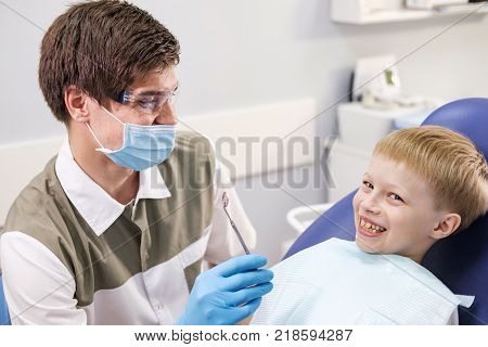 Male dentist examines the teeth of the patient cheerful child with blond hair. Boy smiling in dentist's chair with mouth wide open.