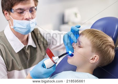 Male dentist examines the teeth of the patient cheerful child with blond hair. Boy smiling in dentist's chair with mouth wide open.