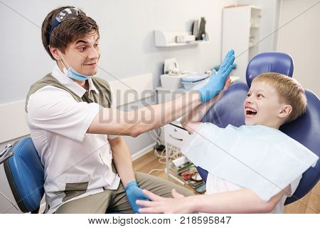 Male dentist examines the teeth of the patient cheerful child with blond hair. Boy smiling in dentist's chair with mouth wide open.