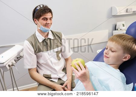 Doctor male dentist giving green apple to patient boy in dental chair
