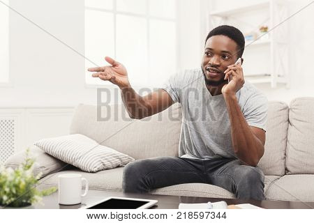 Happy young man talking on smartphone emotionaly sitting on beige couch at home, copy space