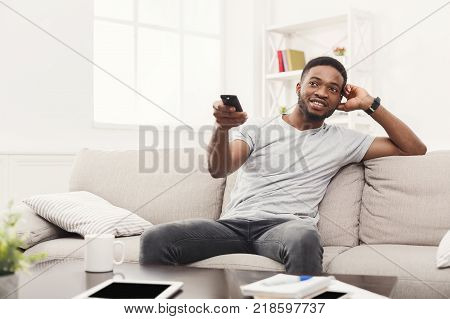 Young happy man watching tv on the couch pointing with remote controller on tv-set, copy space