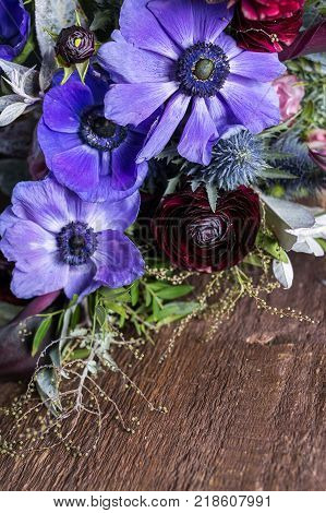growing, floral arrangement, design concept. on the wooden highly textured surface of table there is bouquet of different flowers such as dark like blood battercups and delicate lilac anemons