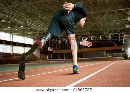 Low angle portrait of determined  amputee sportsman warming up before running practice in modern gym stretching legs, copy space