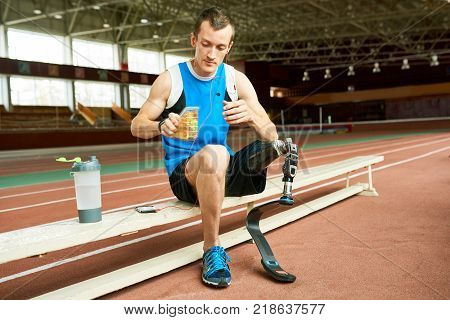 Full length portrait of young amputee athlete taking break from practice on running track and relaxing on bench eating sandwich
