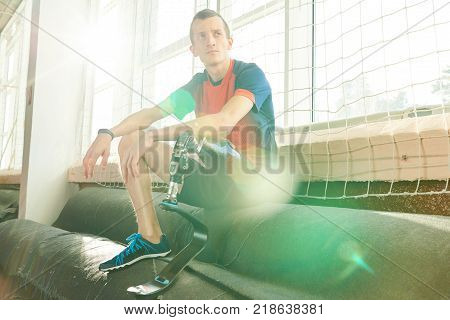 Motivational portrait of young  amputee sportsman sitting on bench in modern gym, focus on prosthetic leg in sunlight with lens flare