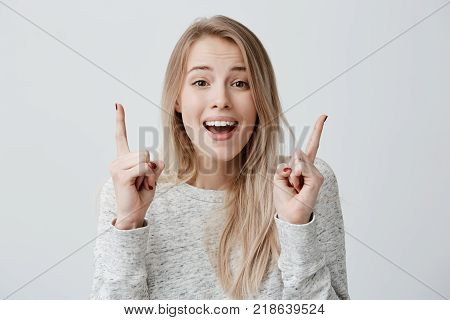 Shot of joyful blonde girl wearing long sleeved t-shirt looking at camera, pointing fingers at copy space above her head. Beautiful young woman smiling indicating something on blank studio wall with hands