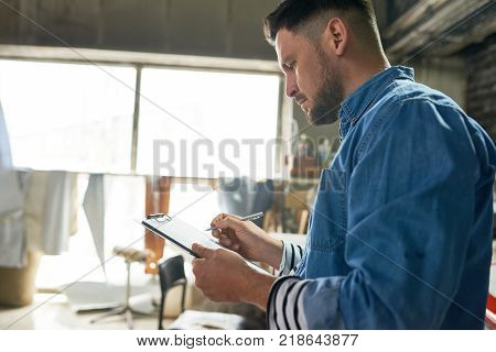 Side view portrait of handsome mature man writing on clipboard while working in crafting workshop, copy space