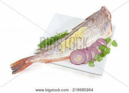 Carcass of the uncooked red cod without head with cilantro and dill twigs lemon sliced red onion on the large square white dish on a white background
