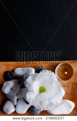 A wooden rectangular board has multiform pebbles, burning aroma candles and a bud of white beautiful flower for spa treatments. On a black background. View from above.