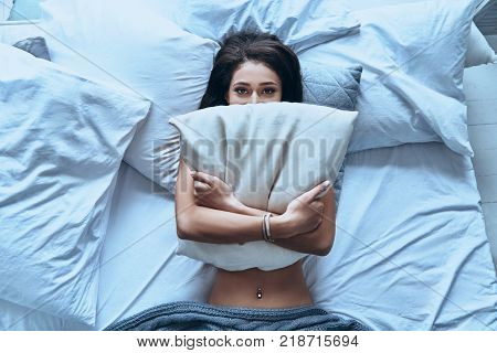 I want to stay in bed. Top view of attractive young woman covering half of her face with pillow and looking at camera while lying on the bed at home