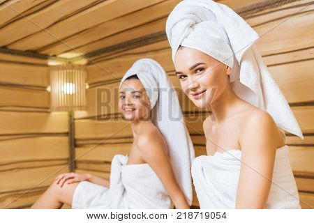 happy young women covered with towels relxing in sauna and looking at camera