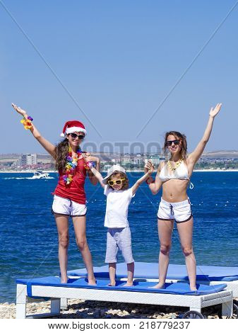 Two sisters and a little brother depict joy, raised their hands up against the background of the sea. One girl in traditional Hawaiian lei. Free space for advertising.