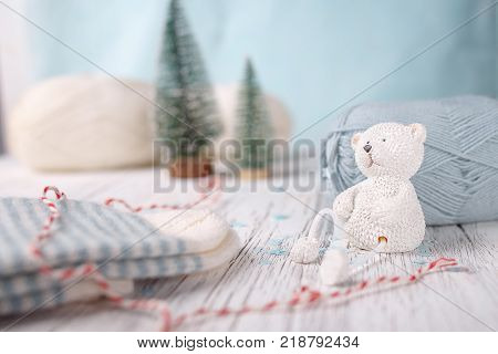 White toy bear sits near woolen light blue ball of yarn. Knitted striped socks, two christmas tree and wool white yarn on white wooden background.