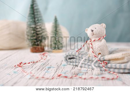 A white toy bear is sitting on a pile of socks. White wooden background on which there are two Christmas trees and a skein of yarn. Selective focus.