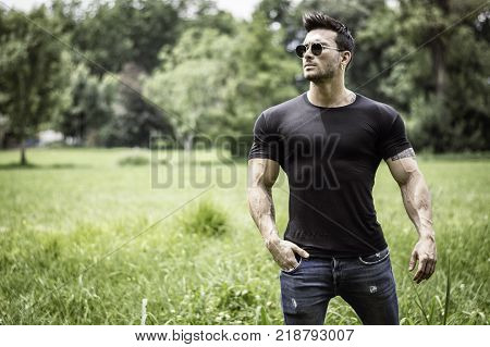 Handsome Muscular Hunk Man Outdoor in City Park or Countryside, Sitting on Grass. Showing Healthy Muscle Body While Looking away