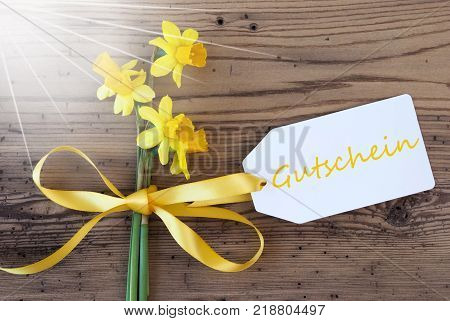 Label With English Text Gutschein Means Voucher. Sunny Yellow Spring Narcissus Or Daffodil With Ribbon. Aged, Rustic Wodden Background. Greeting Card For Spring Season