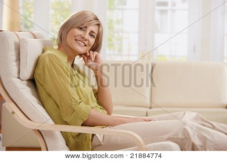 Portrait of adult Woman smiling in die Kamera, die sitzen im Sessel in helles Wohnzimmer mit hand