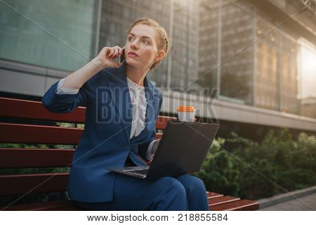 Young woman sitting outdoors and using laptop. Businesswoman working. Holding cup of coffee. and speaking on the phone at the same time.