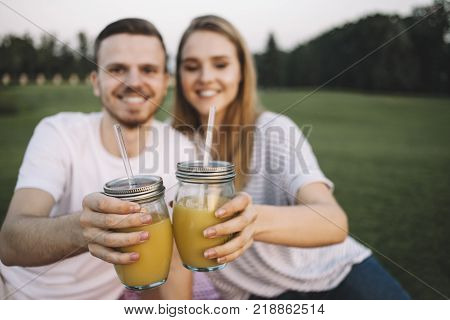 Young couple is sitting in a large gree park together and dringking some juice. The boy is looking straight to the camera while the girl is looking somewhere down. They are enjoying company of one another. Close up. Cut view