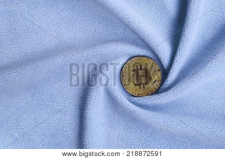 The golden bitcoin lies on a blanket made of soft and fluffy light blue fleece fabric with a large number of relief folds. The shape of the folds resembles a fan from a video card cooler