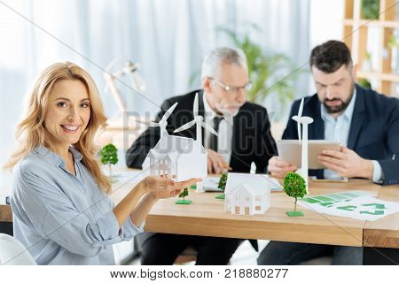 Cheerful woman. Cute emotional friendly woman looking glad and showing a nice model of a house while her enthusiastic colleagues sitting by her side and working at the progressive windmill turbines