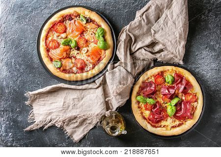 Two Traditional pizza with bresaola, smoked salmon, cheese, tomatoes and basil served on black plates with textile over black texture background. Top view with space.