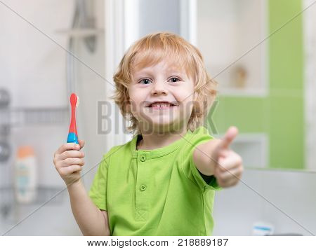 Child boy brushing his teeth in the bathroom. Smiling child holding toothbrush and showing thumbs up.