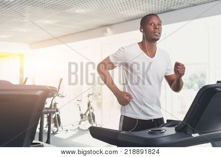 Young happy handsome african-american man in fitness club. Cardio workout, running on treadmill. Healthy lifestyle, guy training in gym. Flare effect, copy space