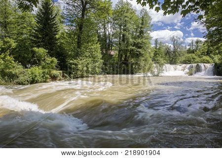 Croatian Village Of Rastoke By A River Canyon With Wooden Houses In A Green Lanscape With Trees And