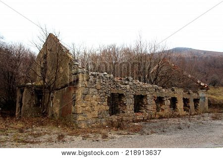 Stone house ruins without roof or windows with trees, plants and other vegetation growing from inside surrounded with gravel, grass and dried trees on cold winter day