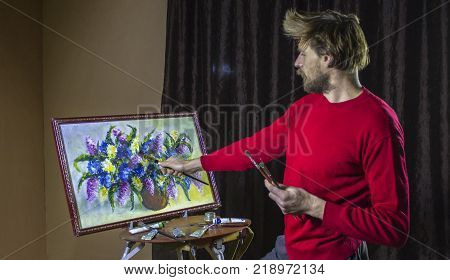 A man bearded artist in a dark sweater draws an artistic brush painting flowers still life on easer in the studio