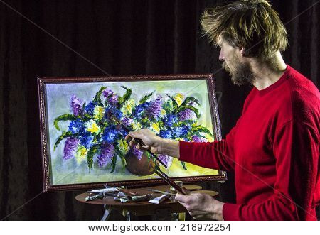 A man bearded artist in a dark sweater draws an artistic brush painting flowers still life on easer in the studio