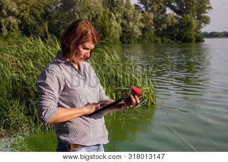 Side View Of Ecologist Entering Data On Green Algae Taken In River On Tablet