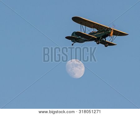 A Green And Yellow Antique Bi Plane Doing A Fly Over Passing A Full Moon With A Blue Sky Background.