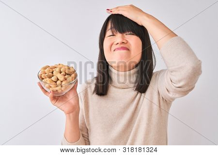 Young beautiful Chinese woman holding bowl with peanuts over isolated white background stressed with hand on head, shocked with shame and surprise face, angry and frustrated. Fear and upset 