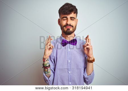 Young man with tattoo wearing purple shirt and bow tie over isolated white background gesturing finger crossed smiling with hope and eyes closed. Luck and superstitious concept.