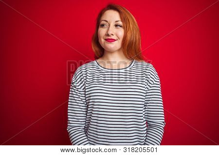 Young redhead woman wearing strapes navy shirt standing over red isolated background smiling looking to the side and staring away thinking.